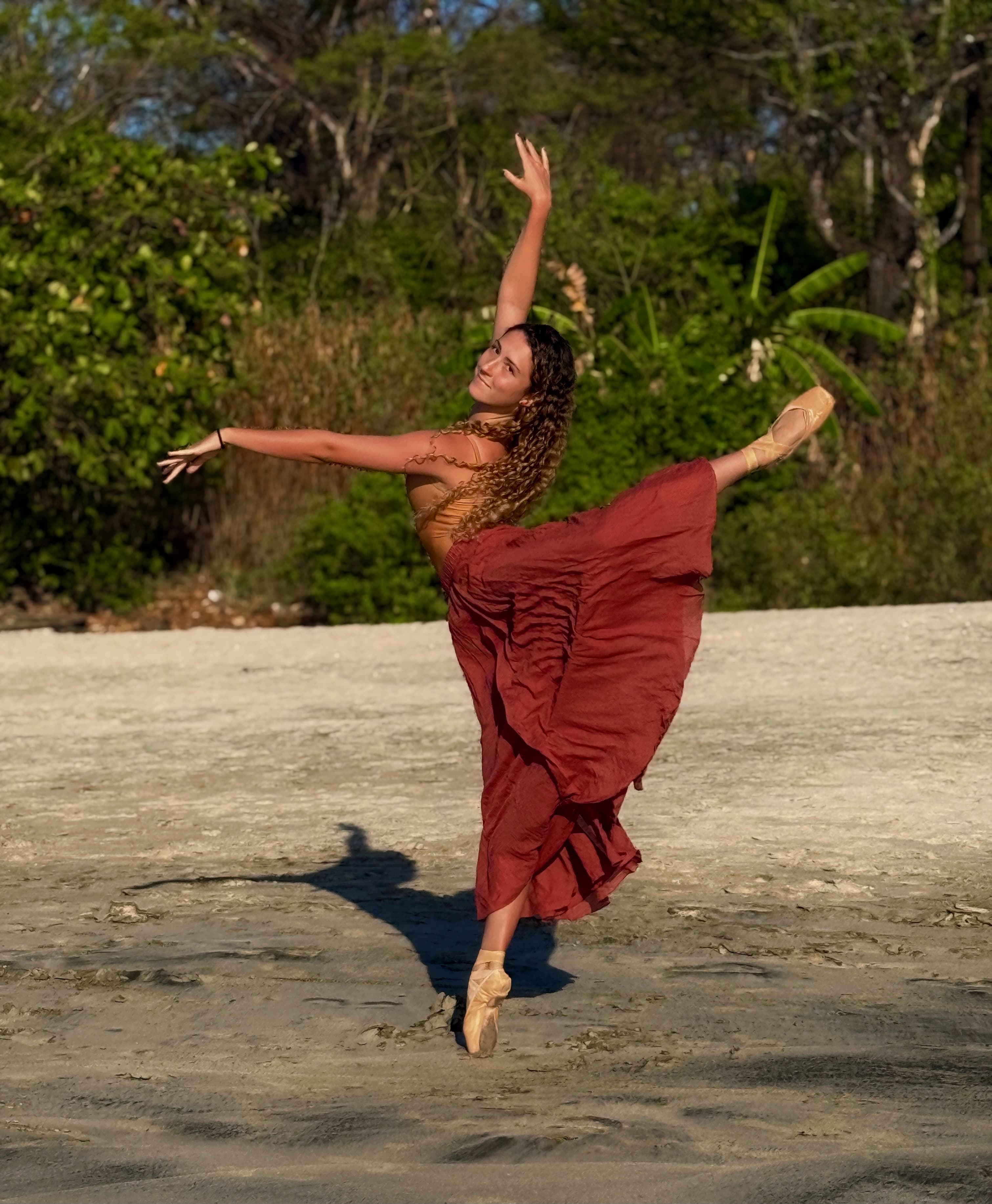 A ballerina in a flowing red skirt performs a graceful pose on a sandy beach.